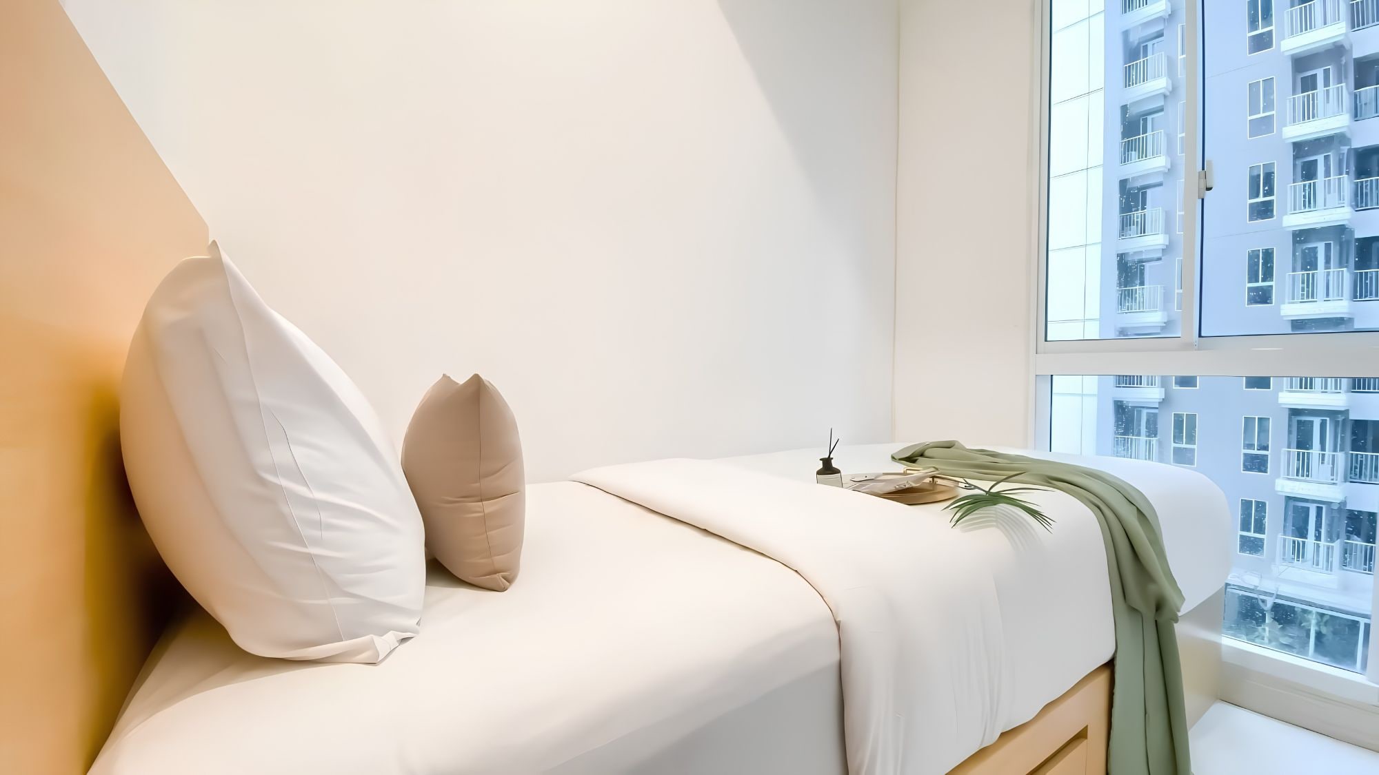 A minimalist bedroom with a bed, pillows, a bedside table, a plant, and a view of a building through a window.