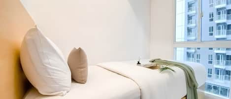 A minimalist bedroom with a bed, pillows, a bedside table, a plant, and a view of a building through a window.