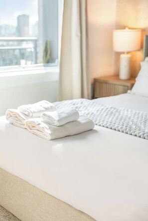 A neatly made bed with folded white towels, a bedside lamp, and a view of a cityscape through a window. - The Selena - Luxury Condo w/ Rooftop Pool, Gym (Orlando)