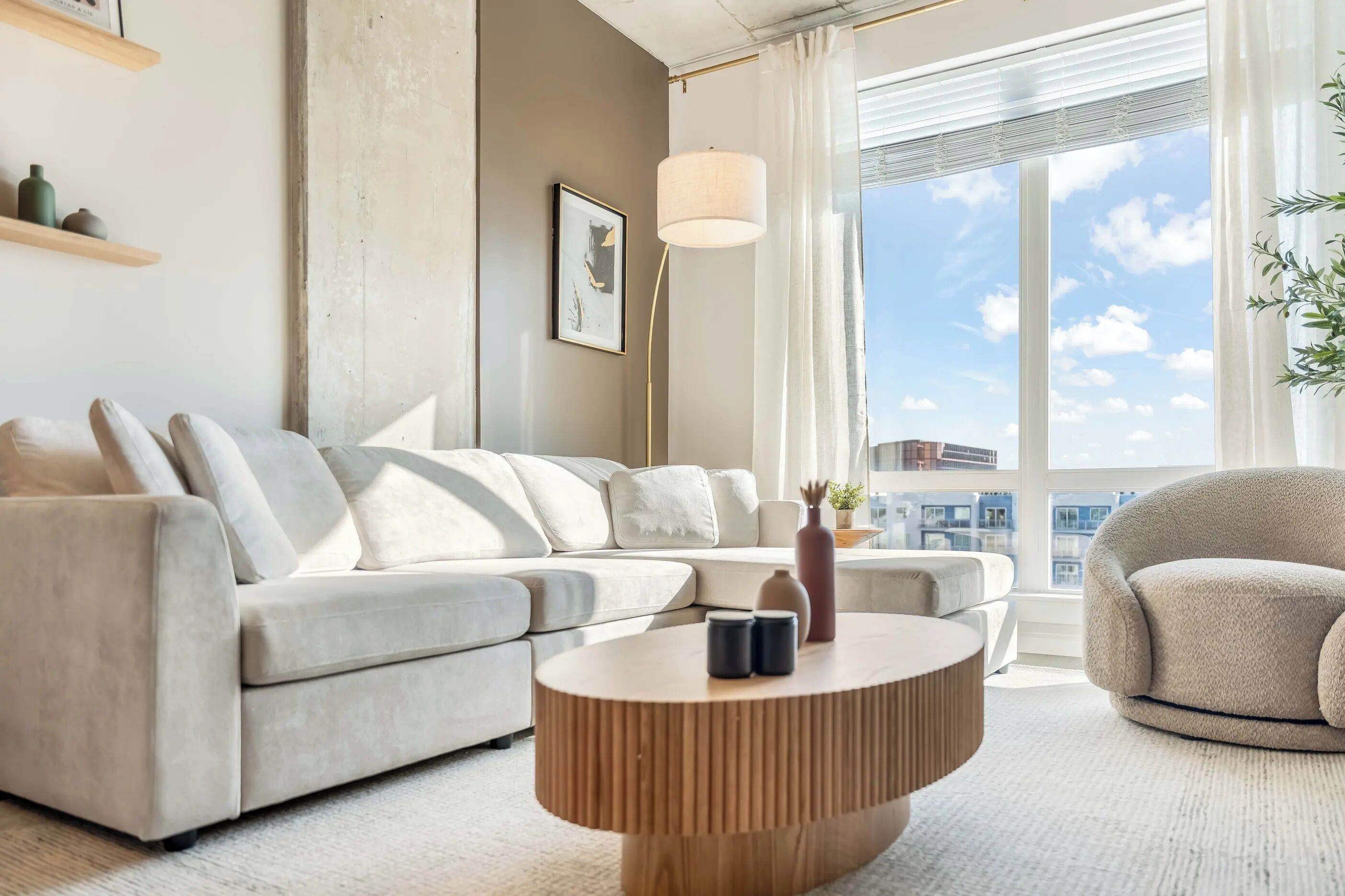 Ein modernes Wohnzimmer mit einem grauen Sofa, einem runden Couchtisch und Blick auf die Stadt durch große Fenster.