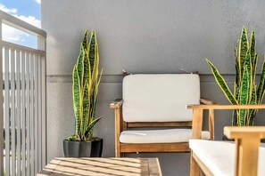 Ein Balkon mit einem Holzstuhl, einem kleinen Tisch, zwei Kübelpflanzen und einem weißen Geländer.