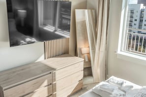 Ein Schlafzimmer mit einem Bett, einer Holzkommode, einem flachbildfernseher an der Wand und einem Fenster mit Vorhängen.