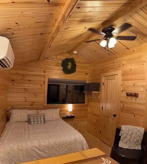 A wooden cabin room with a bed, a ceiling fan, a wall-mounted heater, and a window with a wreath.