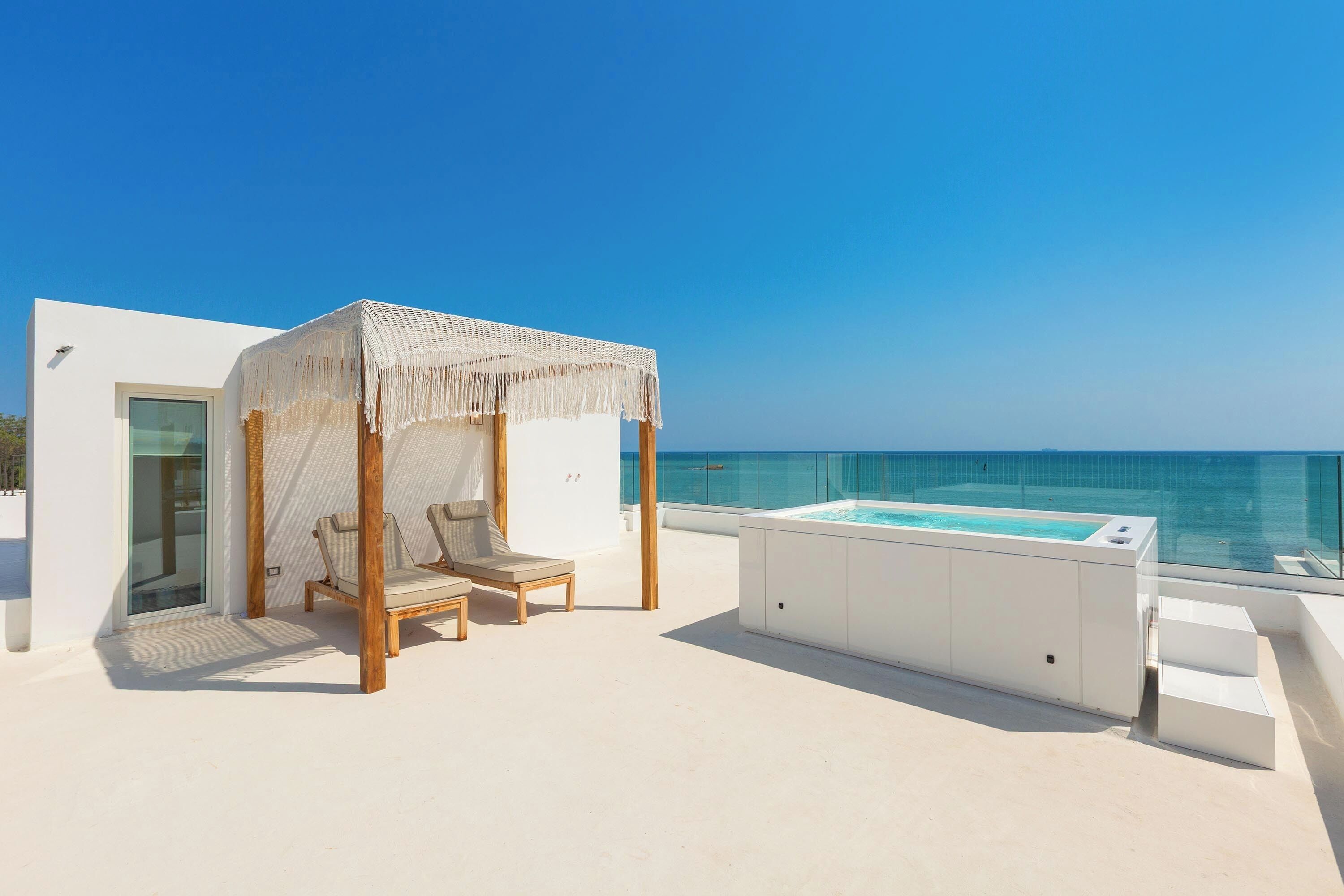A rooftop terrace with a glass balustrade, a hot tub, and two lounge chairs under a woven canopy.