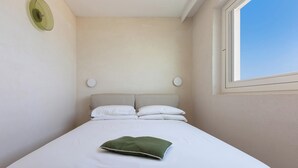 A single bed with white bedding and a green pillow, a round wall clock, and a window with a clear view of the sky.