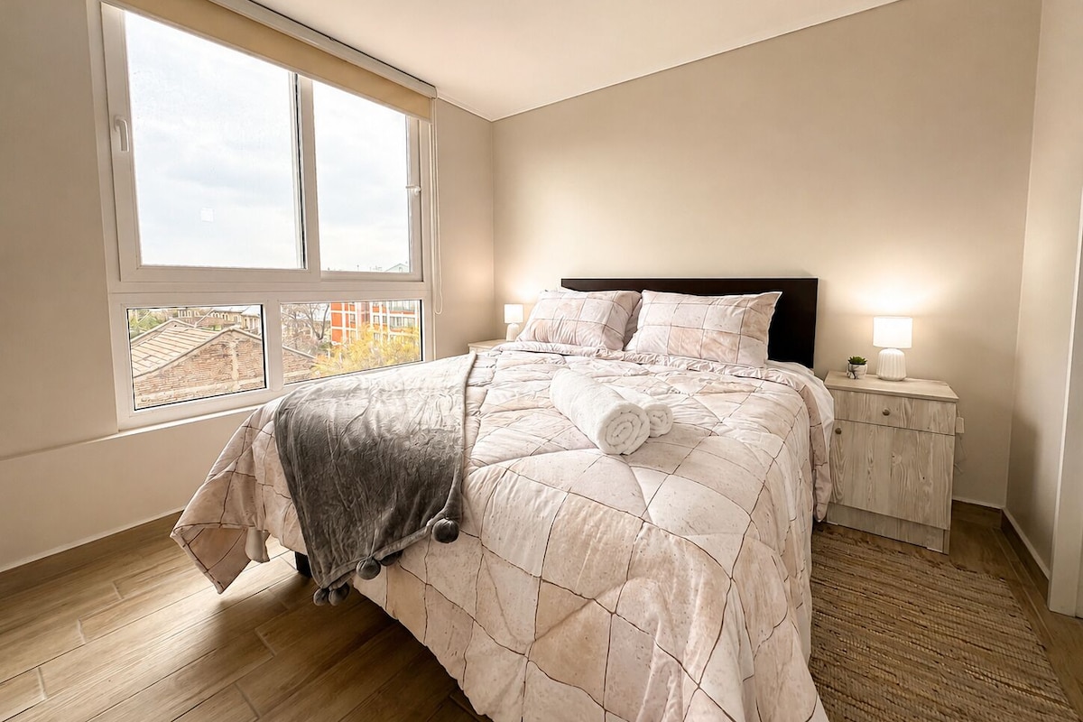 A bedroom with a large window, a bed with a quilt, a bedside table with a lamp, and a nightstand with a plant.