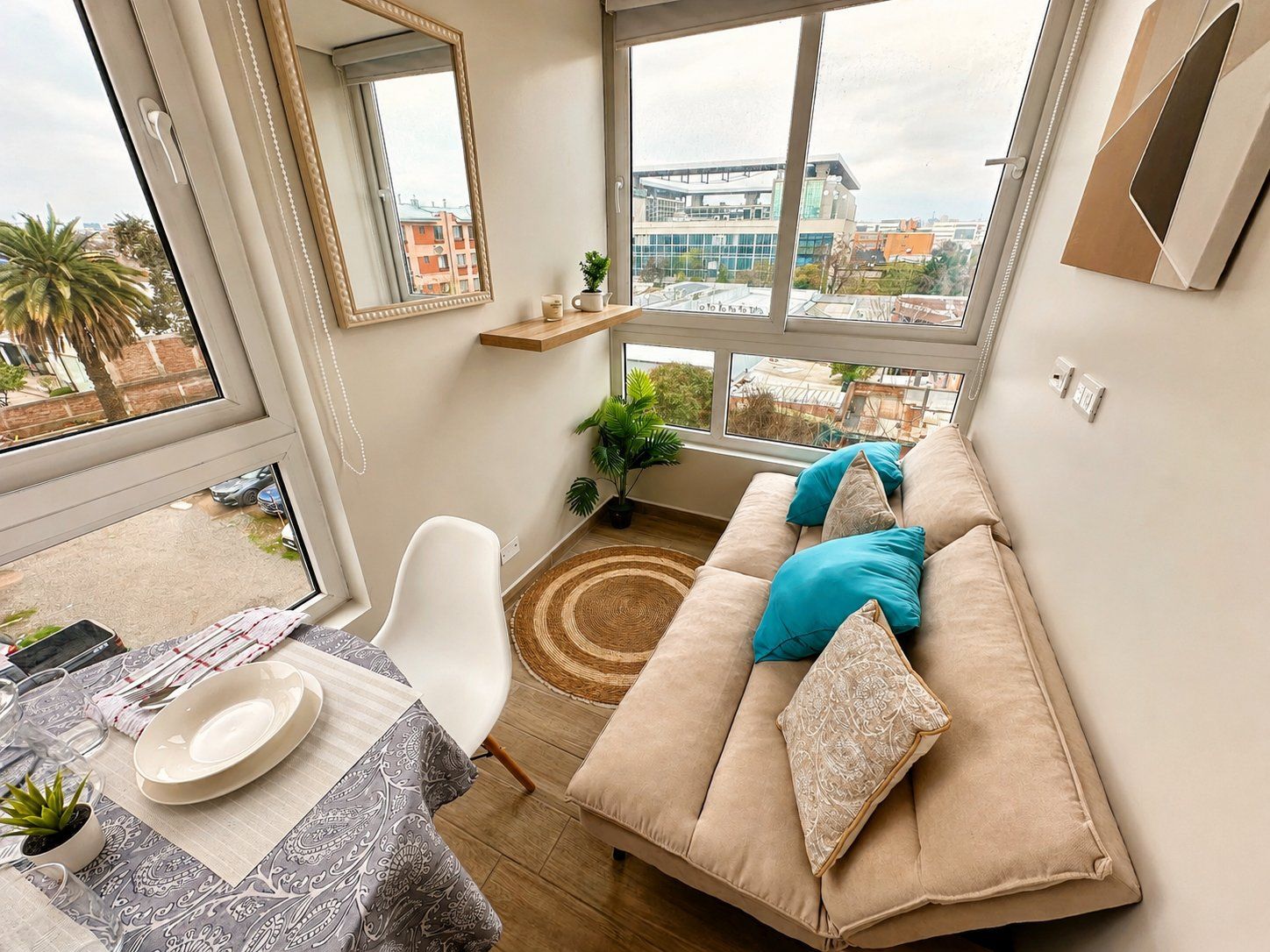 A modern living room with a sofa, a dining table with plates and glasses, a mirror, and a view of buildings through large windows.