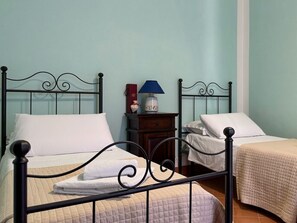 Habitación con dos camas, cada una con una almohada blanca y una toalla blanca doblada. Hay una mesita de noche con una lámpara y un objeto decorativo.