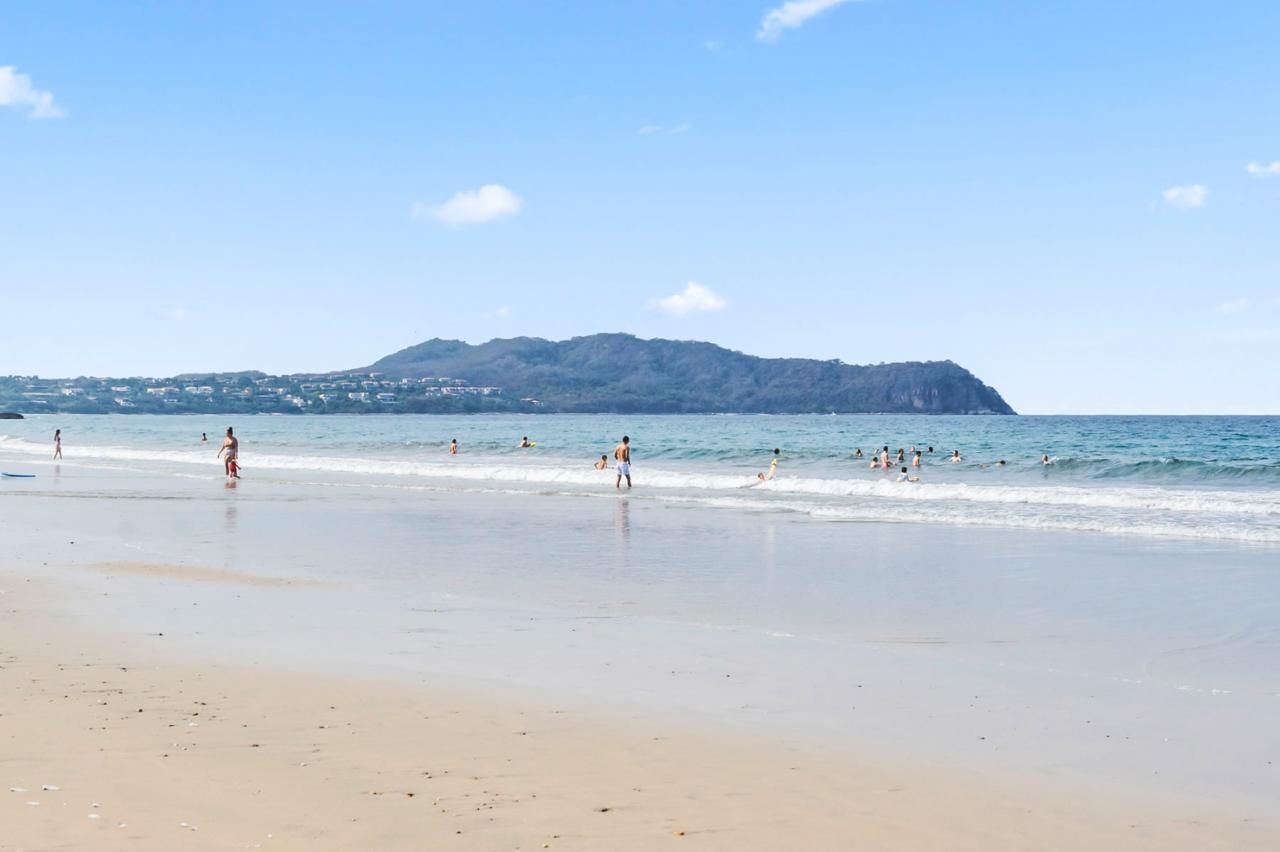 Una playa con gente nadando y caminando por la orilla, una montaña al fondo y un cielo despejado.
