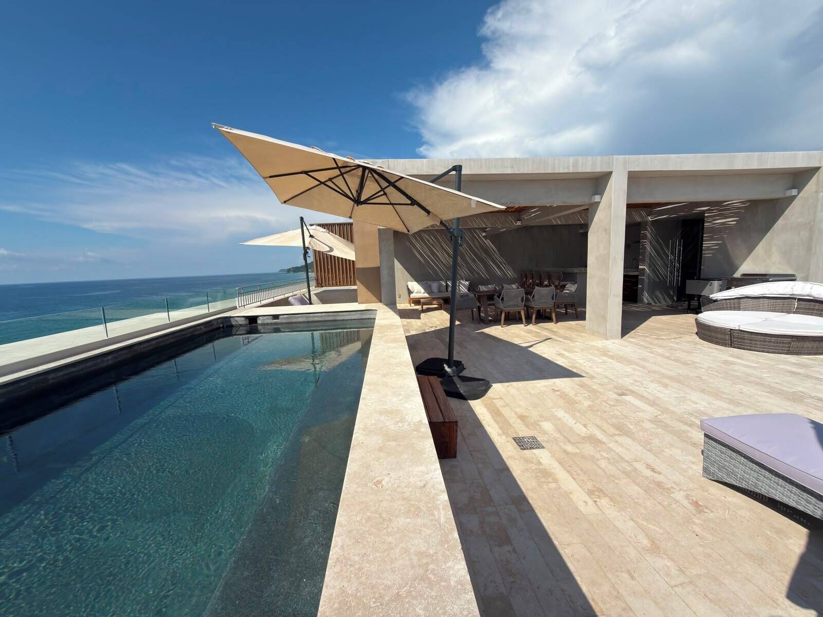 Ein moderner Poolbereich im Freien mit freiem Blick auf den Ozean, eine Sitzgelegenheit mit Tischen und Stühlen sowie ein großer Sonnenschirm, der Schatten spendet.