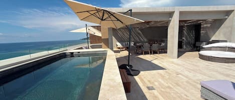 A modern outdoor pool area with a clear view of the ocean, a seating area with tables and chairs, and a large umbrella providing shade.