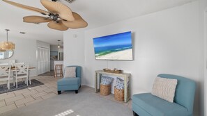 A living room with a ceiling fan, a fireplace, a beach-themed painting, and a dining area with a table and chairs. - Coastal Retreat- steps from the beach, pool included (Gulf Shores)