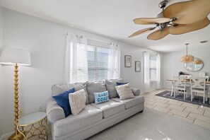 A modern living room with a grey sofa, a floor lamp, and a ceiling fan. - Coastal Retreat- steps from the beach, pool included (Gulf Shores)