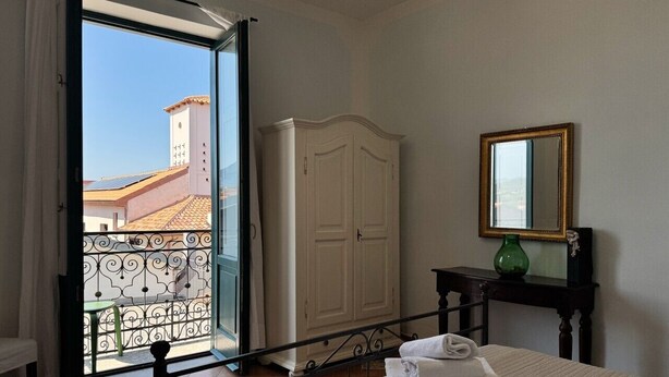 A hotel room with a bed, a wardrobe, a small table with a vase, and a balcony with a view of buildings.