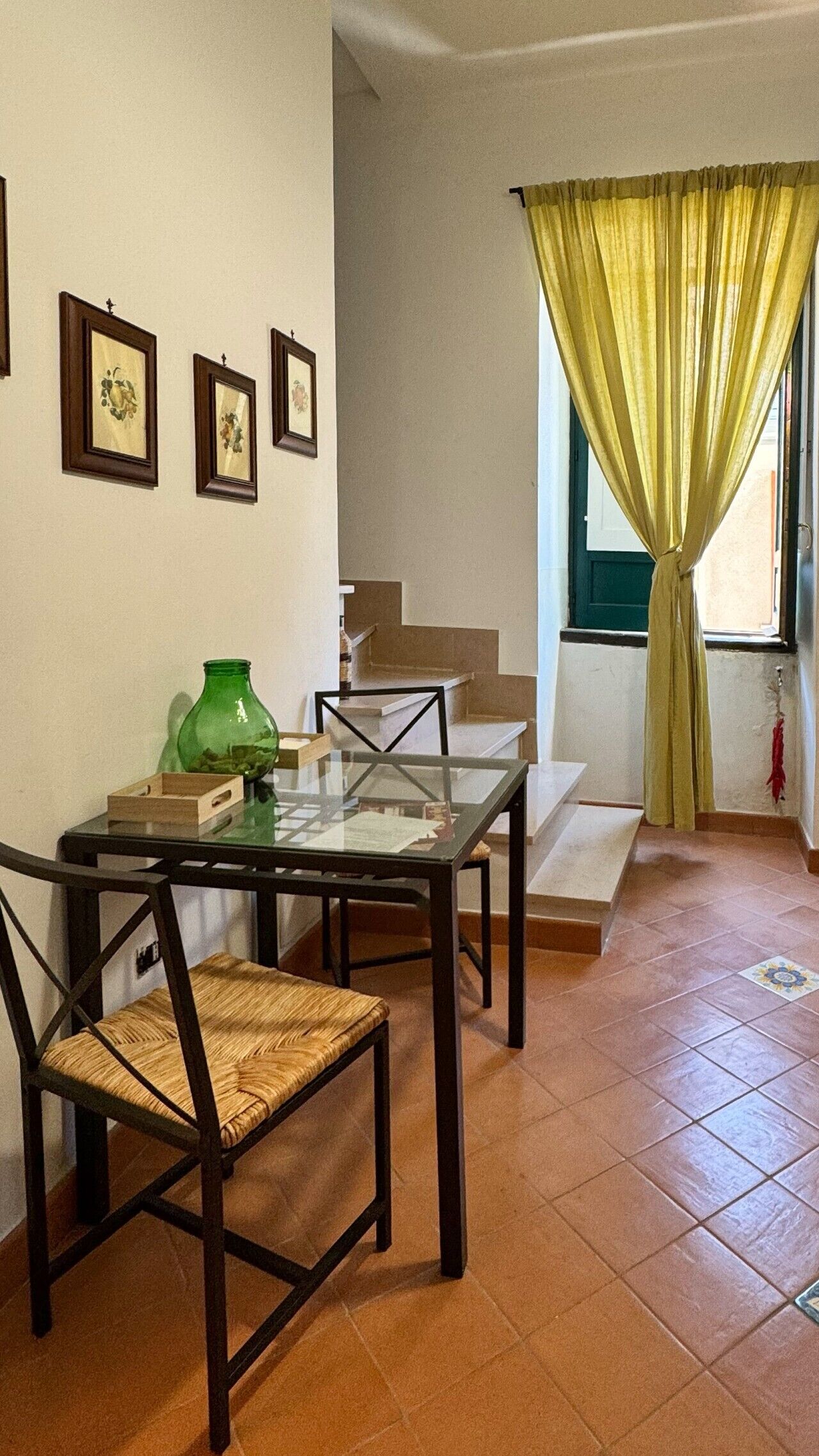 Uma área de refeições com uma mesa de vidro, duas cadeiras, uma garrafa verde e quadros na parede.