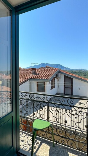 Un balcón con barandilla de metal, una silla verde y vistas a edificios con tejados rojos y montañas al fondo.