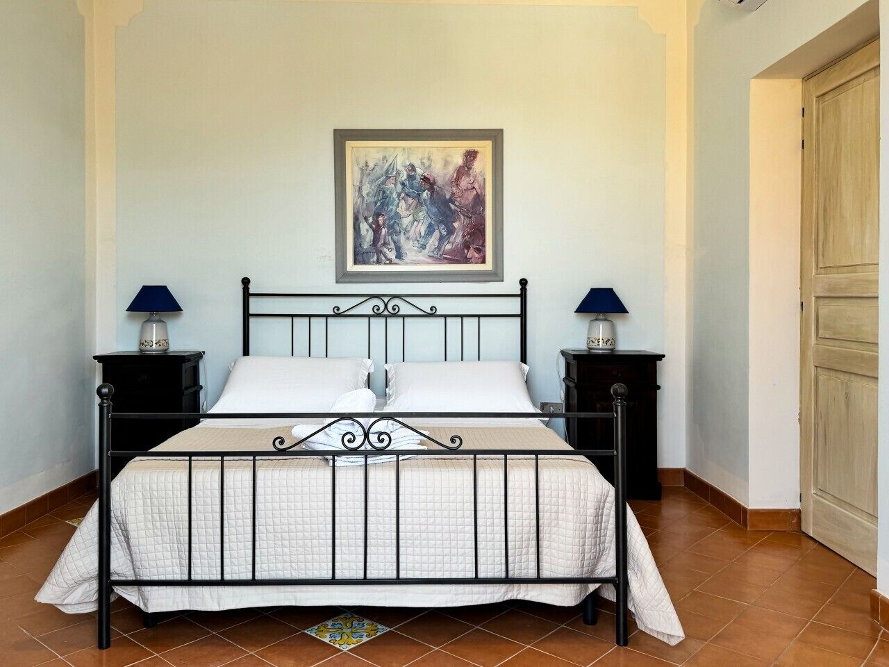 A bed with a metal frame, two bedside tables with lamps, a painting on the wall, and a wooden door.