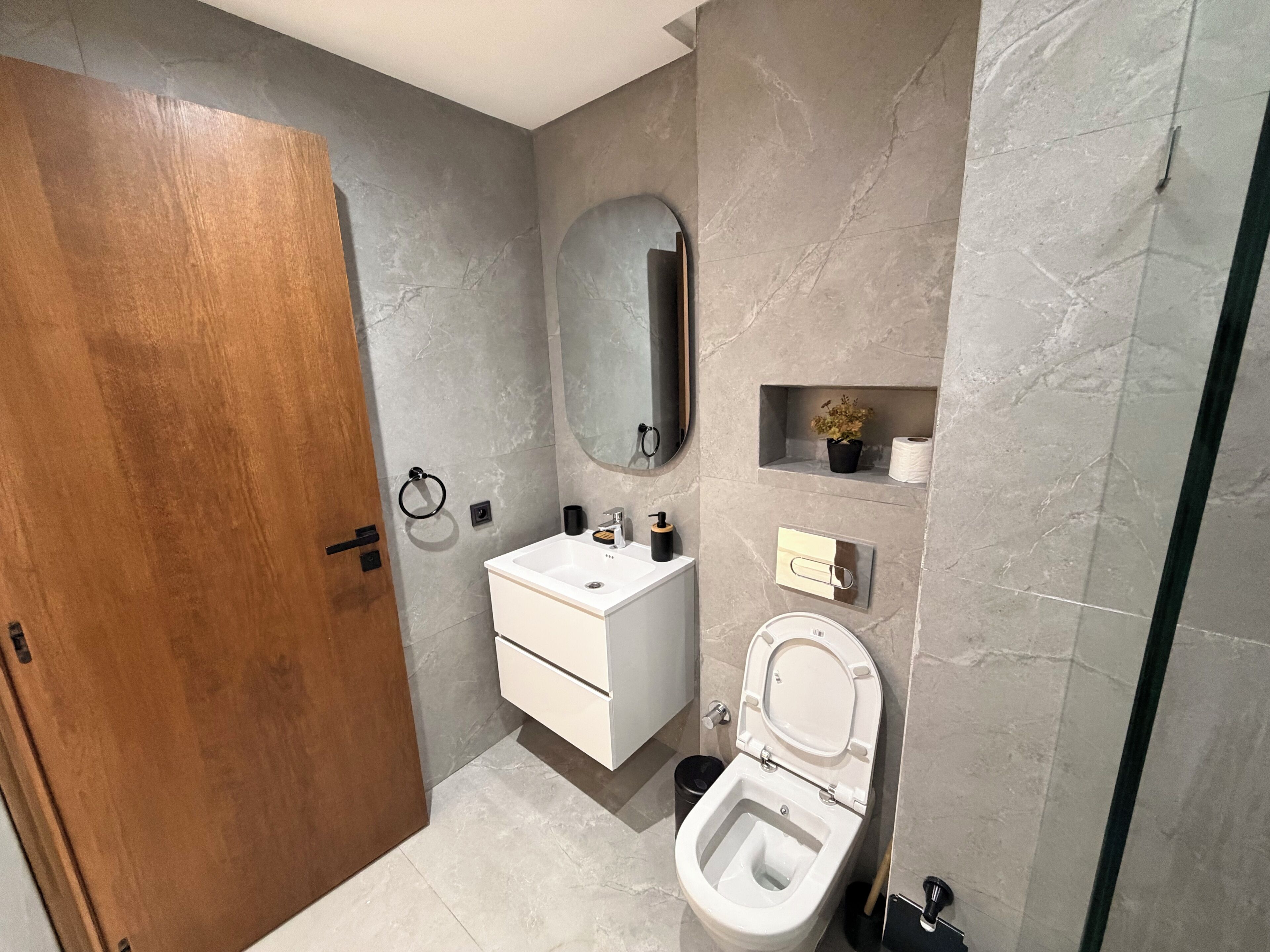 Un baño moderno con puerta de madera, un lavabo blanco, un inodoro y un espejo.