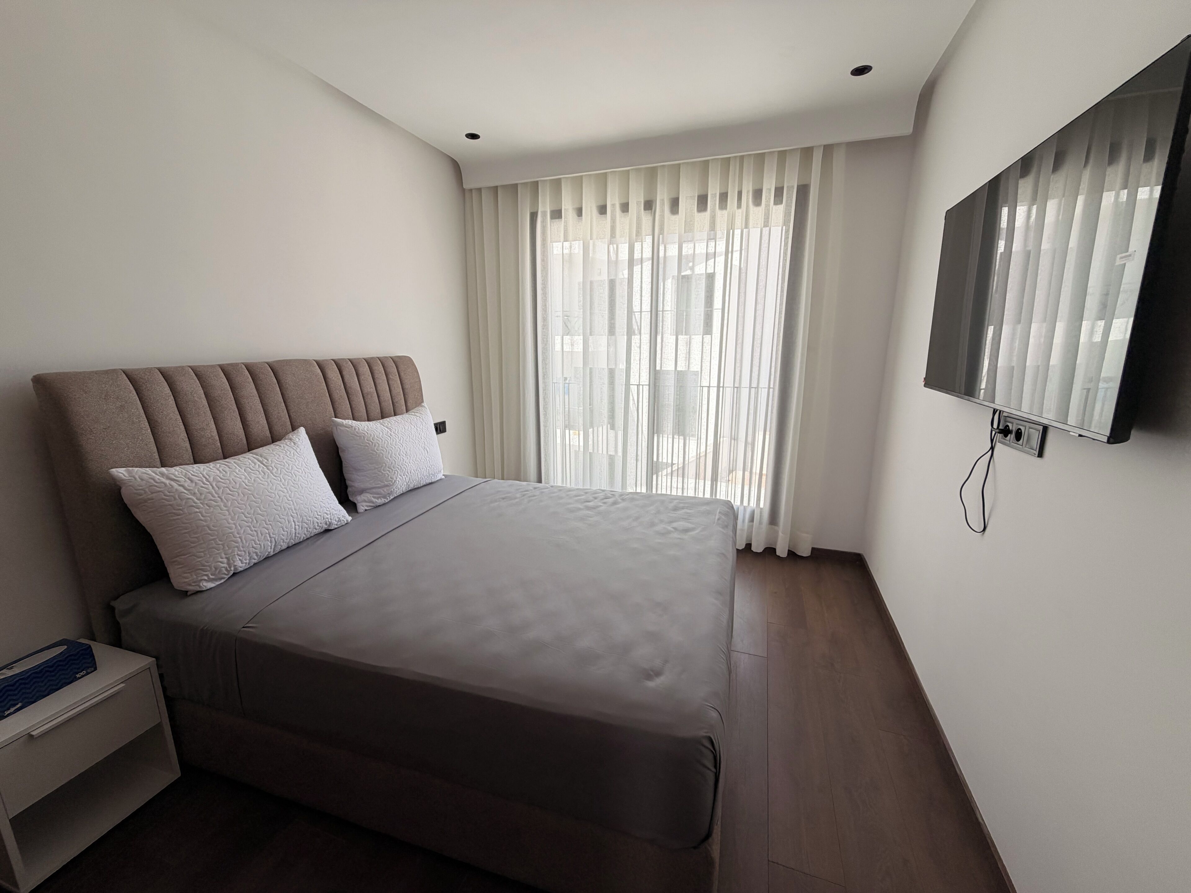 Un dormitorio moderno con una cama grande, una mesita de noche, un televisor montado en la pared y una ventana con cortinas traslúcidas.