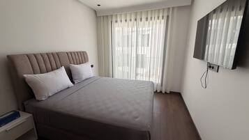 Un dormitorio moderno con una cama grande, una mesita de noche, un televisor montado en la pared y una ventana con cortinas traslúcidas.