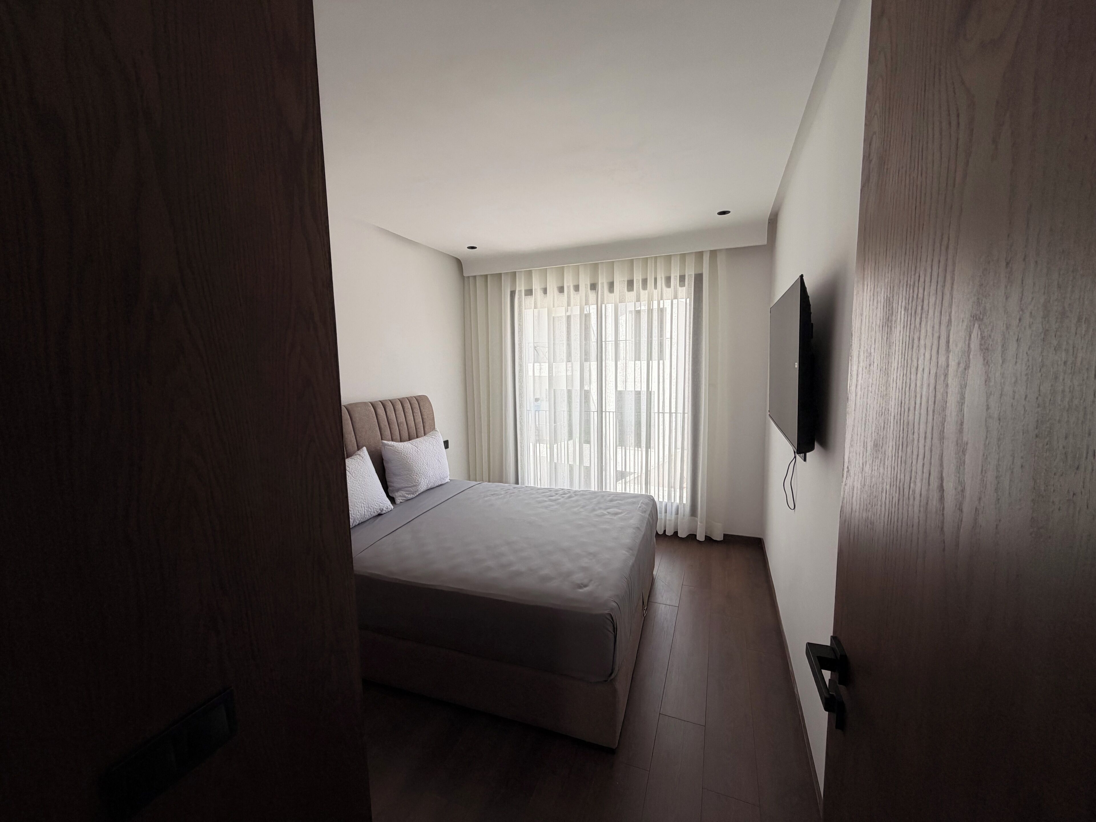 Un dormitorio con cama, televisor en la pared y ventana con cortinas.