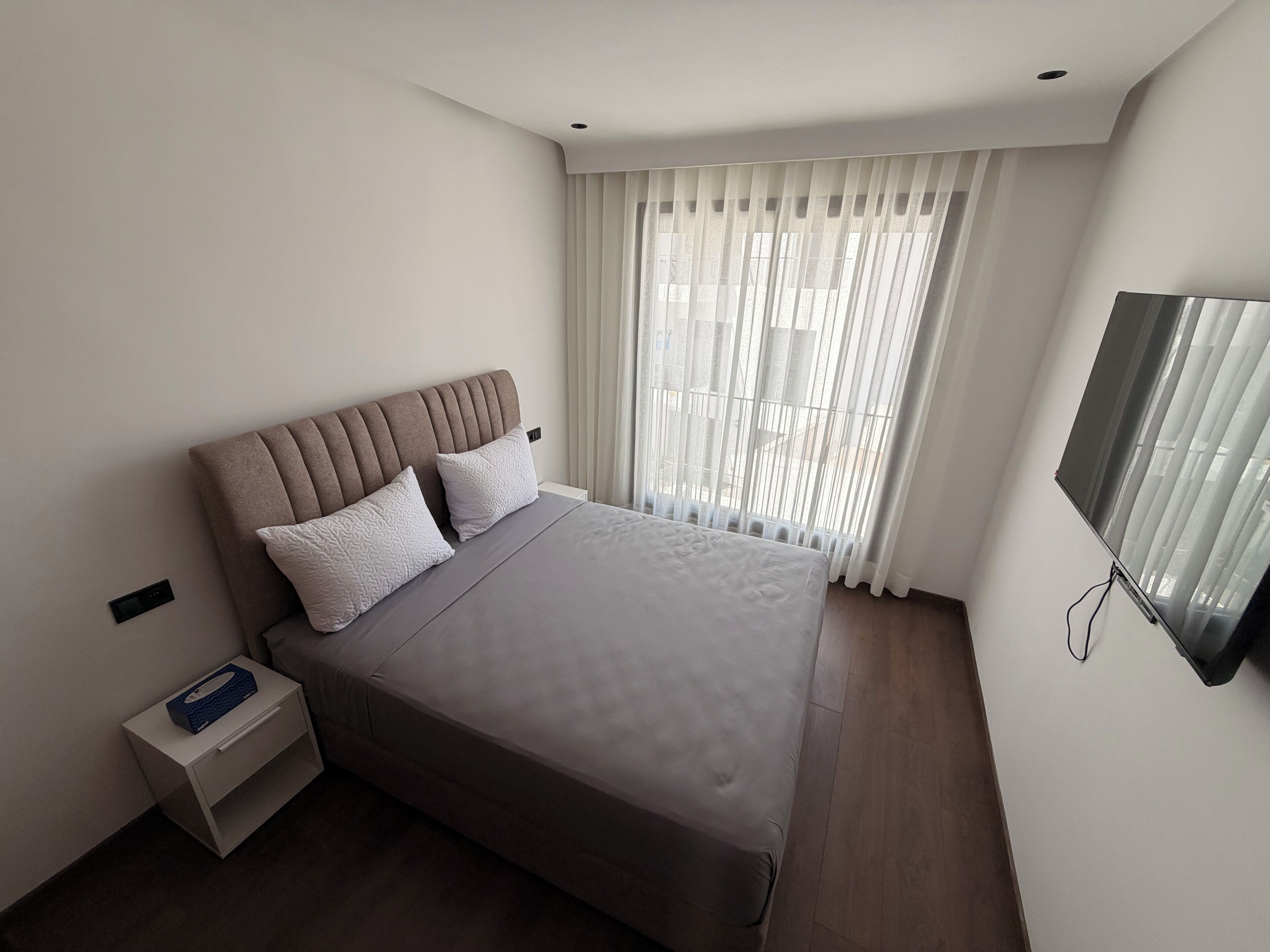 A modern bedroom with a bed, a nightstand, a television, and a window with curtains.