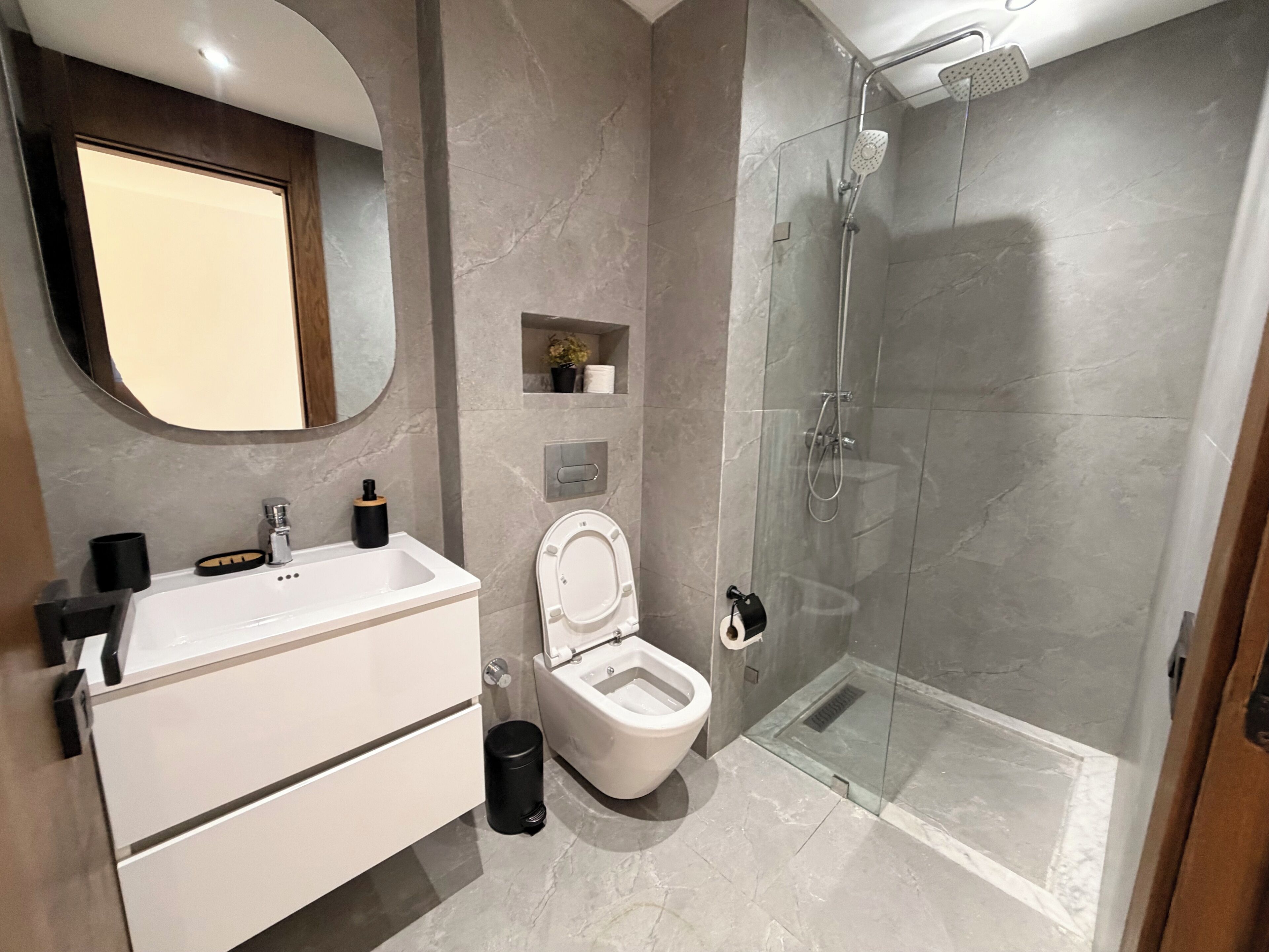 A modern bathroom with a white sink, mirror, and toilet, a glass-enclosed shower, and a small shelf with toiletries.