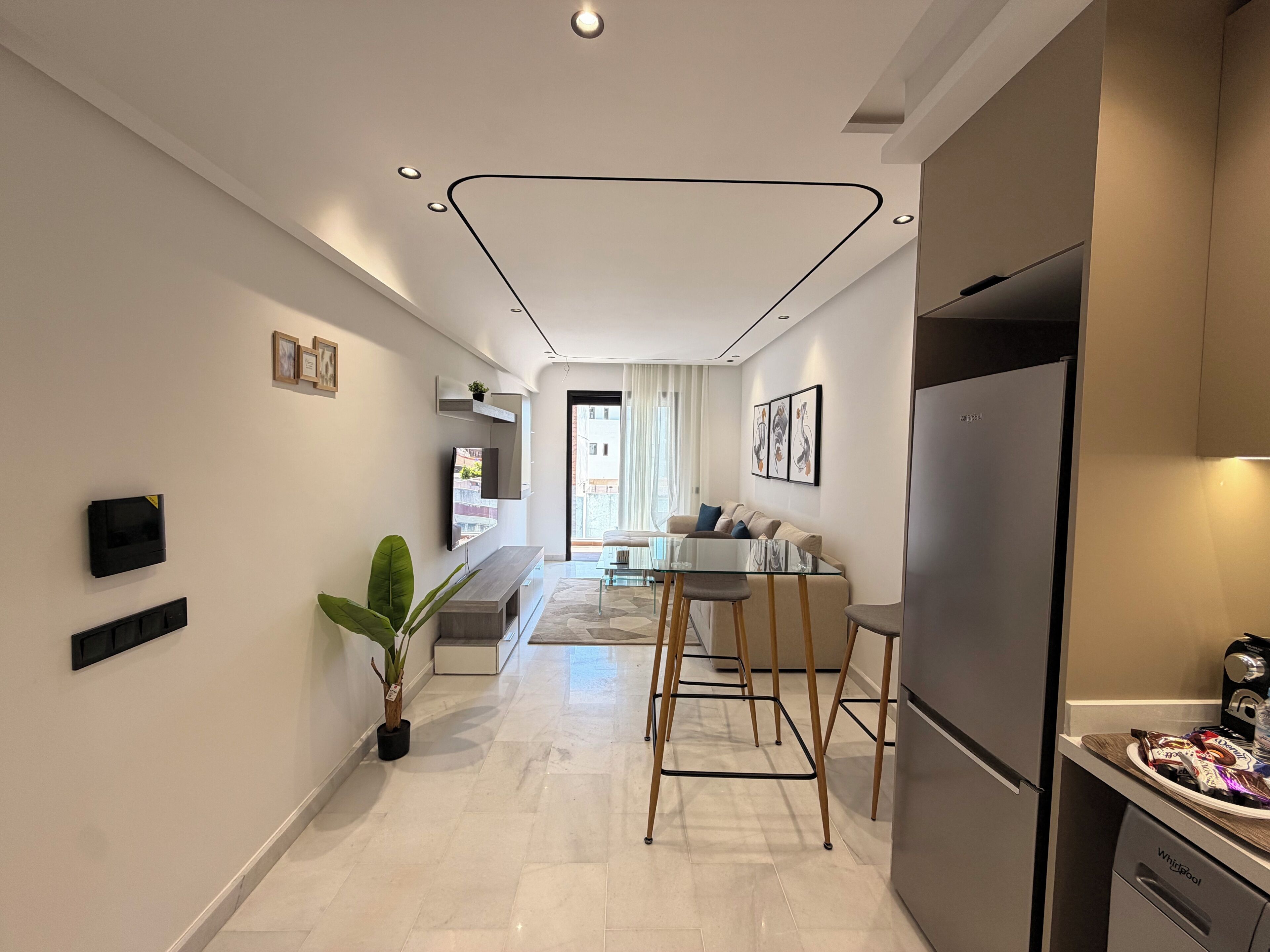 A modern kitchen with a refrigerator, a dining area with a glass table and chairs, and a living area with a sofa and a TV mounted on the wall.