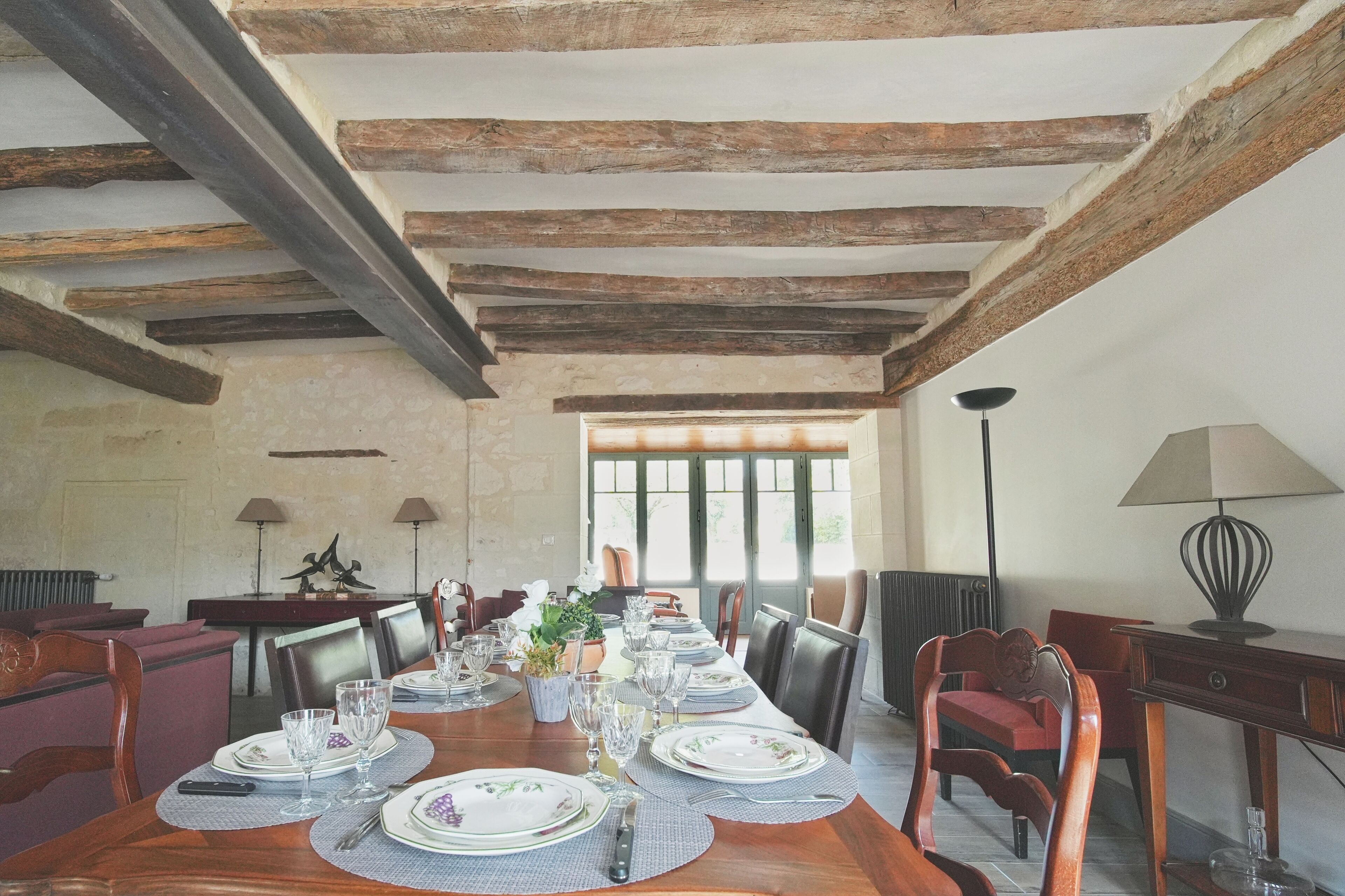 A dining room with a long wooden table set for a meal, flanked by chairs, with a rustic ceiling and a view through a window.