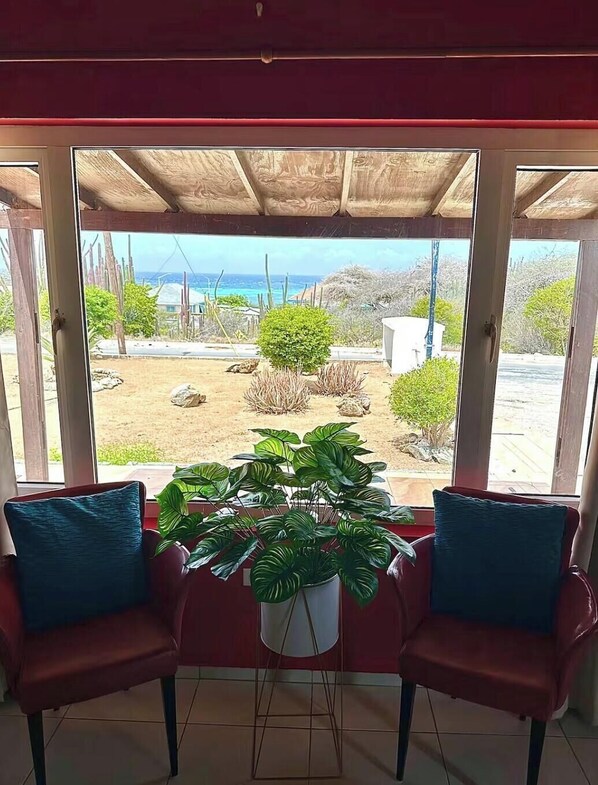 A room with a view of a garden and a beach. There are two chairs with cushions and a potted plant in the center.
