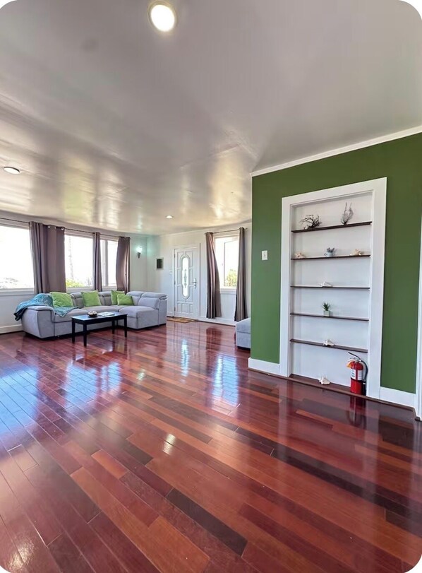 A spacious living room with hardwood flooring, a grey sofa, and a white shelving unit against a green wall.