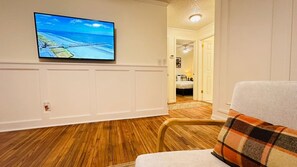 A room with a television mounted on the wall, a wooden floor, a chair with a plaid cushion, and a door leading to another room.