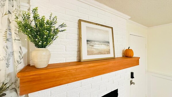A white brick fireplace with a wooden mantel, a framed picture, a potted plant, and a pumpkin.