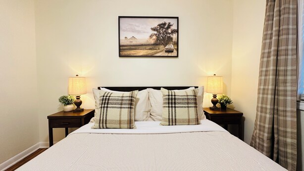 A bed with a headboard, two bedside tables with lamps, a framed picture on the wall, and a window with curtains.