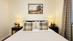 Een bed met hoofdbord, twee nachtkastjes met lampen, een lijst met een afbeelding aan de muur, en een raam met gordijnen.