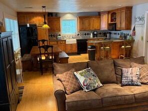 A cozy living room with a brown sofa, a coffee table, and a kitchen area with wooden cabinets and a tiled backsplash. - Emerald Point On Grand (Jay)