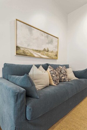 Un sofà blau amb diversos coixins adossat a una paret sense decoració, amb un quadre emmarcat a la part superior.