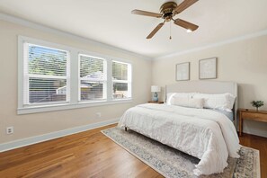 Une chambre à coucher comprenant un lit, une table de chevet, une lampe, un ventilateur de plafond et de grandes fenêtres équipées de stores.
