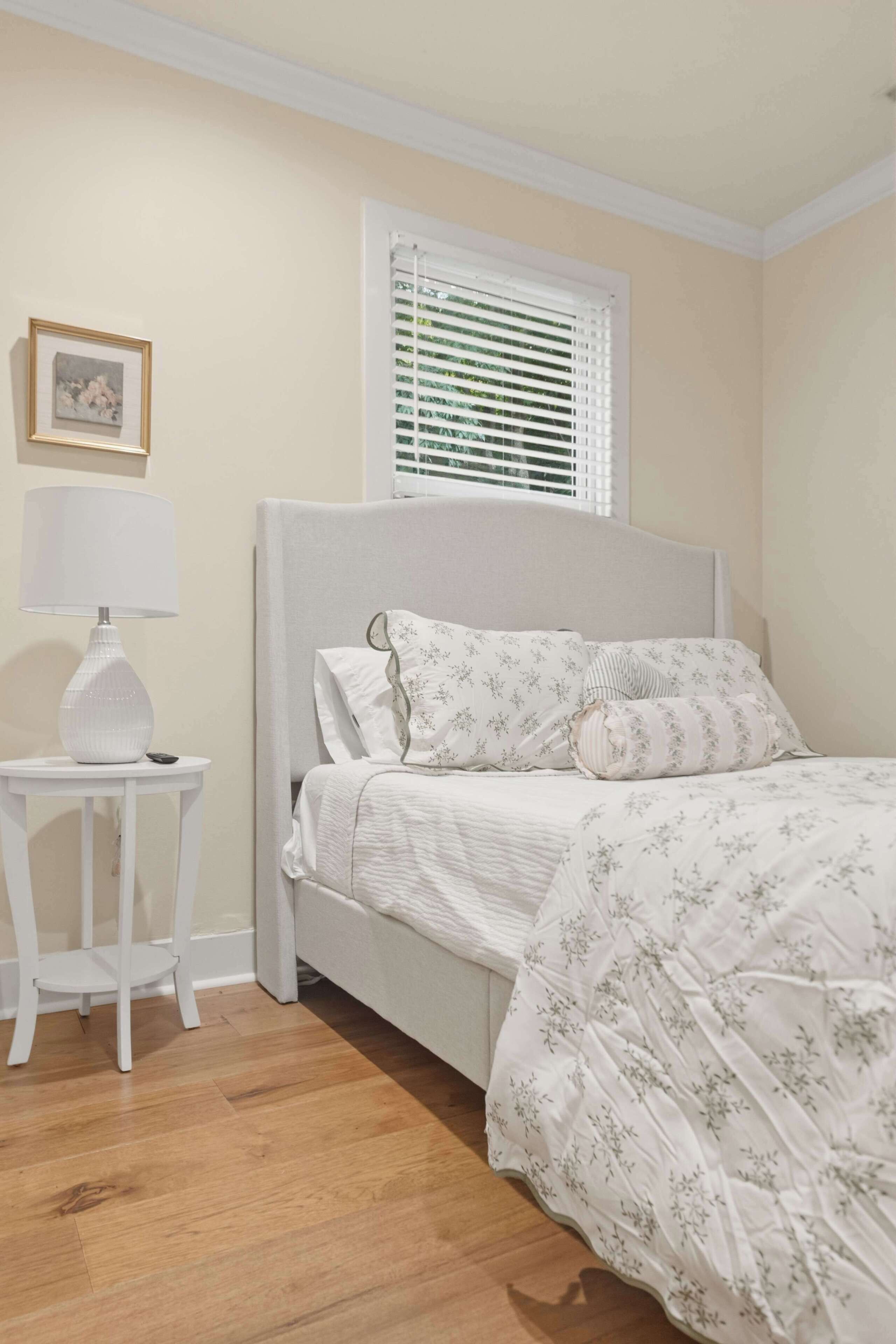 A bedroom with a bed, a bedside table with a lamp, a window with blinds, and a framed picture on the wall.