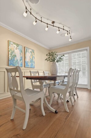 Une salle à manger avec une table en bois et des chaises blanches, un vase avec des plantes vertes et deux tableaux accrochés au mur.