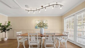 Une salle à manger avec une table en bois, des chaises blanches, un vase rempli de fleurs et deux tableaux accrochés au mur.