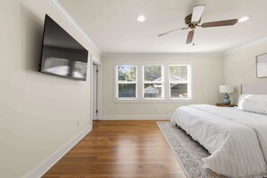 Un dormitorio con cama, ventilador de techo, televisor montado en la pared y ventana con persianas.