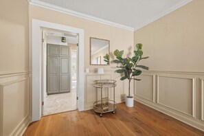 Un pasillo con suelo de madera, una planta en maceta, un espejo y una puerta que da a otra habitación.