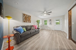 A spacious living room with a dark leather sofa, a yellow floor lamp, a wooden clock, and a ceiling fan.