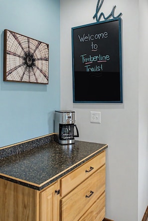 A kitchen with a coffee maker on the counter, a wall clock, and a welcome sign.