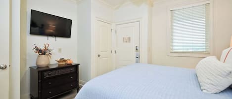 Un dormitorio con cama, cómoda, televisor, ventilador de techo y ventana con persianas.