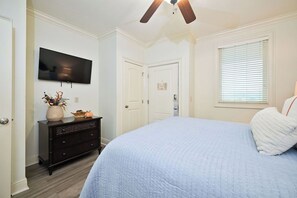Een slaapkamer met een bed, een dressoir, een televisie, een plafondventilator en een raam met jaloezieën.