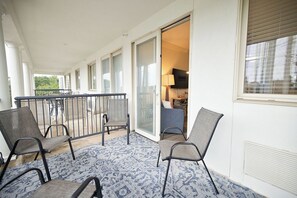 Een balkon met stoelen en een tapijraad, een glazen schuifdeur die leidt naar een kamer met een televisie, en een witte buitenmuur.