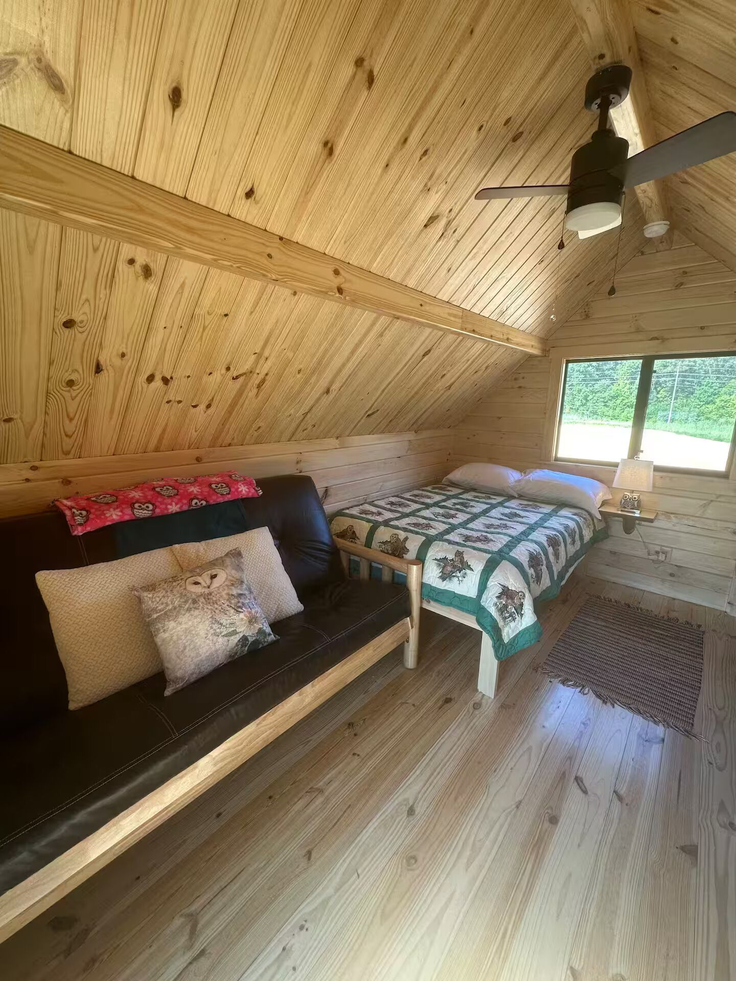 Interior acogedor de una cabaña con techo de madera, una cama con colcha verde y blanca, un sofá de cuero negro con cojines estampados y una mesita con una lámpara.