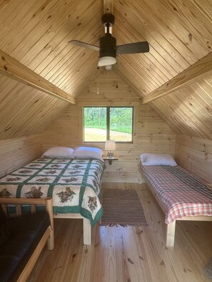 A cozy attic bedroom with two beds, a ceiling fan, and a window providing a view of the outdoors. - Lakehouse Resort Hootin’ Hollow A-Pod 4 Rend Lake (Benton)